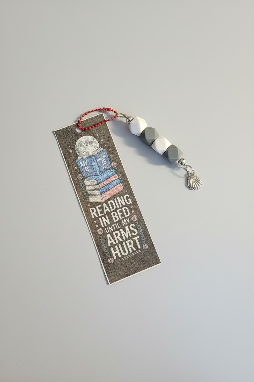 Bookmark with text 'Reading in bed until my arms hurt' on a dark surface with blurred text.